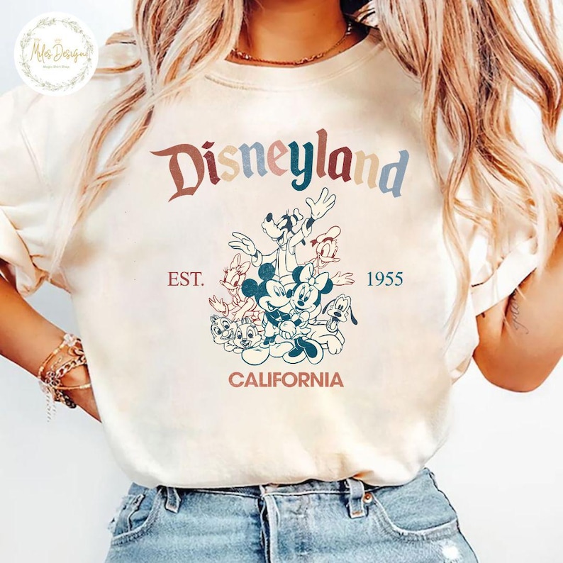 May include: A white t-shirt featuring a vintage-style graphic of Mickey Mouse and his friends in front of the Disneyland logo. The text reads "Disneyland EST. 1955 CALIFORNIA".