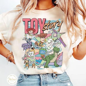 Pode incluir: Uma t-shirt de cor creme com uma impress&atilde;o gr&aacute;fica de personagens de Toy Story, incluindo Jessie, Woody, Buzz Lightyear, Slinky, Rex e outros. O design inclui o texto TOY story, SINCE 1995 e nomes de personagens num layout de estilo retro.