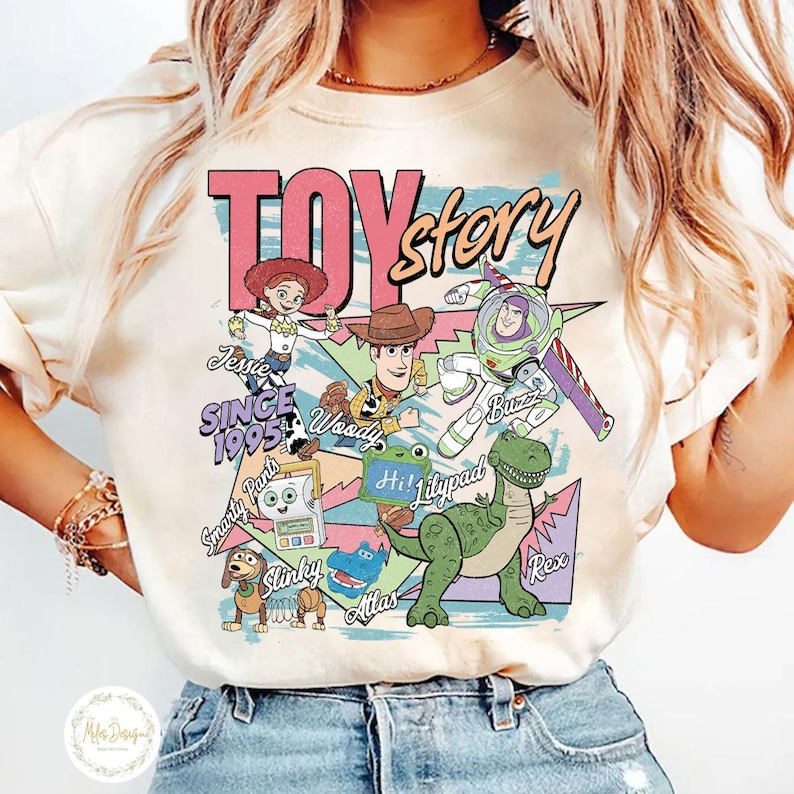 Pode incluir: Uma t-shirt de cor creme com uma impress&atilde;o gr&aacute;fica de personagens de Toy Story, incluindo Jessie, Woody, Buzz Lightyear, Slinky, Rex e outros. O design inclui o texto TOY story, SINCE 1995 e nomes de personagens num layout de estilo retro.