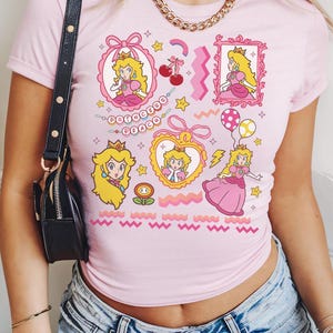 May include: Pink t-shirt featuring multiple cartoon images of Princess Peach, with the text "Princess Peach". The design includes stars, cherries, and a rainbow. The shirt is a crew neck and short-sleeved.