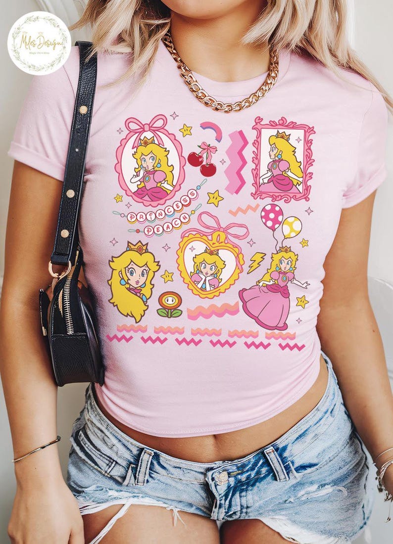 May include: Pink t-shirt featuring multiple cartoon images of Princess Peach, with the text "Princess Peach". The design includes stars, cherries, and a rainbow. The shirt is a crew neck and short-sleeved.