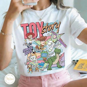 Pode incluir: Uma t-shirt branca de manga curta com uma impress&atilde;o gr&aacute;fica colorida de personagens de Toy Story, incluindo Jessie, Woody, Buzz Lightyear, Rex e Slinky. O texto diz Toy Story, Since 1995, Jessie, Woody, Buzz, Smarty Pants, Lilypad, Slinky e Rex.