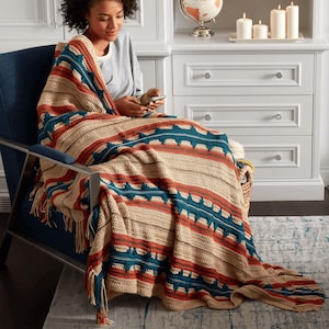 May include: A crocheted throw blanket with a geometric pattern in shades of brown, orange, and teal. The blanket has fringe on the edges and is draped over a chair.