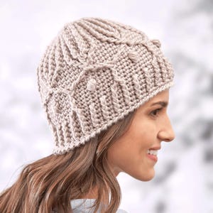 May include: A beige crocheted beanie with a cable knit pattern. The beanie is worn by a person with long brown hair.