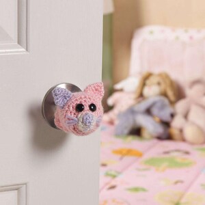 May include: A pink and purple crocheted cat door knob cover. The cat has black eyes and a pink nose.
