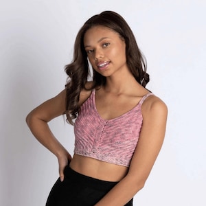 May include: A pink knitted crop top with a V-neck and thin straps. The top has a textured, ribbed design and a light pink and gray color scheme. The top is paired with a black bottom.