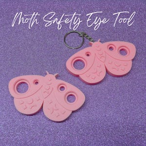 May include: Two pink moth-shaped safety eye tools with keychains. The tools have circular openings and are decorated with wing details. The text "Moth Safety Eye Tool" is written in white script above the tools. The background is a sparkly purple.