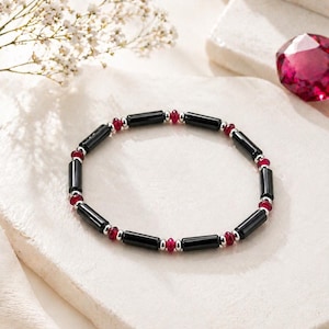 May include: A black beaded bracelet with red accents. The bracelet features alternating black, rectangular beads and small, round red beads. The bracelet is displayed on a white surface with a flower and a large, faceted red gemstone in the background.