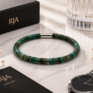 May include: A green and silver beaded bracelet with a magnetic clasp. The bracelet is displayed with a black box and a watch. The box has the letters "RJA" printed on it.