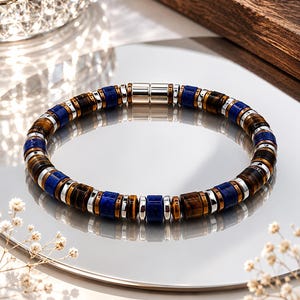 May include: A beaded bracelet featuring alternating cylindrical tiger eye beads in shades of brown, deep blue lapis lazuli beads, and small silver-toned metallic spacers. The bracelet is secured with a polished silver-toned cylindrical magnetic clasp.