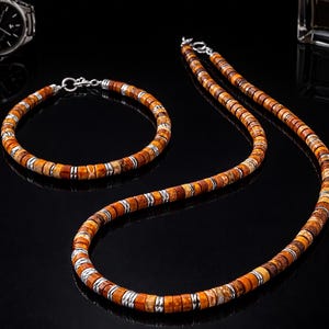 Puede incluir: Un conjunto de joyas con un collar y una pulsera con cuentas marrones y naranjas separadas por detalles metálicos plateados. Un reloj de pulsera plateado y un frasco de perfume son visibles en el fondo, todo sobre una superficie negra.