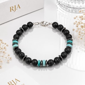 May include: A black lava stone bracelet with turquoise and silver-tone accents. The bracelet features round, porous black beads interspersed with turquoise and silver-tone spacers. The clasp is silver-tone. The bracelet is displayed on a white surface.