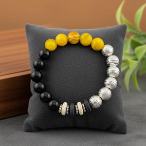May include: A beaded bracelet featuring black, yellow, and silver beads. The bracelet has a mix of round and textured beads, with a central section of smaller, contrasting beads. The bracelet is displayed on a gray cushion.