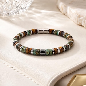 May include: A beaded bracelet featuring alternating disc-shaped tiger eye stones, turquoise-colored stones with dark matrix patterns, and small silver-toned metal spacers. The bracelet includes a cylindrical dark grey bead and a silver-toned clasp.