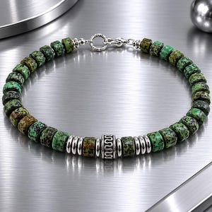 May include: A beaded bracelet featuring green and brown stone beads interspersed with silver-colored metal accents. The bracelet has a decorative central bead and a clasp closure. The jewelry is displayed on a metallic surface.