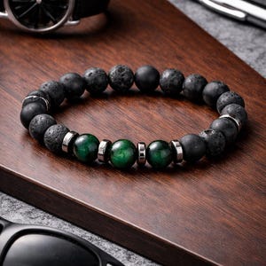 May include: A beaded bracelet featuring black lava stone beads and green gemstone beads, accented with silver-tone metal spacers. The bracelet is displayed on a wooden surface, alongside a black watch and sunglasses.