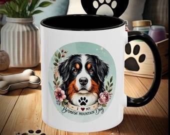 Peut inclure: Une tasse en céramique blanche avec une anse et un bord noirs. La tasse présente une illustration colorée d'un chien de montagne bernois avec des fleurs et le texte "J'aime mon chien de montagne bernois".