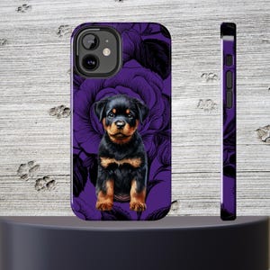 May include: Purple phone case with a black and brown Rottweiler puppy sitting in the center. The background is a purple floral pattern.