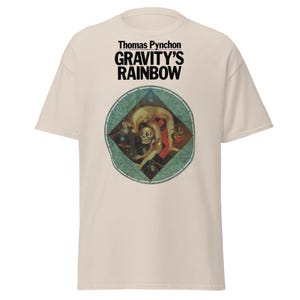 May include: A cream-colored t-shirt featuring the text "Thomas Pynchon GRAVITY'S RAINBOW" in black. Below the text is a circular graphic with a figure in a contorted pose, with a skull head, set against a teal and red background.