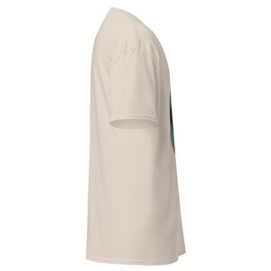 May include: A side view of a cream-colored t-shirt. The short-sleeved shirt is made of a soft material. The shirt has a crew neck and a relaxed fit. The shirt is a solid color with a graphic on the front.