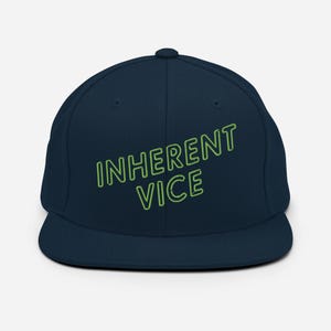 May include: A navy blue baseball cap with a green outline of the text "INHERENT VICE" on the front.