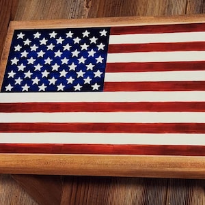 May include: A framed wooden sign featuring the American flag. The flag design includes a blue field with white stars and red and white stripes. The frame is made of light-colored wood.