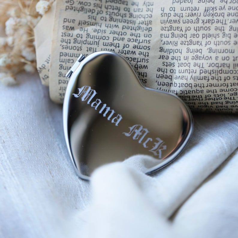 May include: A heart-shaped, silver compact mirror with the engraved text "Mama McK" in a gothic font. The mirror is resting on a white cloth, with a backdrop of a vintage newspaper.