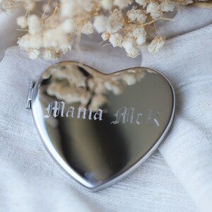 May include: A heart-shaped, silver-colored compact mirror with the engraved text "Mama McR" in a gothic font. The mirror is on a white cloth, with dried flowers in the background.