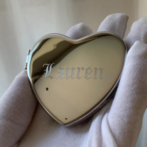 May include: A heart-shaped, silver-colored compact mirror. The name "Lauren" is engraved in an elegant font on the front. The compact is held by a gloved hand.