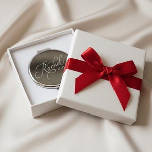 May include: A silver compact mirror engraved with the name "Rachel" and the date "12.21" inside a white gift box. The box is tied with a red satin ribbon and bow. The background is a soft, off-white fabric.