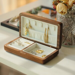 May include: Open wooden jewelry box with a light-colored interior. The box contains various gold and silver jewelry pieces, including earrings, necklaces, rings, and charms. The box is open, revealing the contents. The wood has a warm, natural tone.