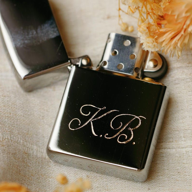 K&ouml;nnte beinhalten: Ein schwarzes, rechteckiges Feuerzeug mit den Initialen "K.B." in kursiver Schrift graviert. Das Feuerzeug ist ge&ouml;ffnet und zeigt sein silbernes Innere. Getrocknete Blumen sind im Hintergrund.