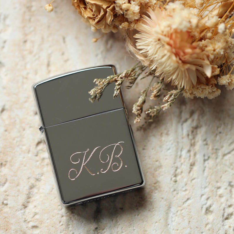 K&ouml;nnte beinhalten: Ein silberfarbenes Feuerzeug mit den eingravierten Initialen "K.B." in Schreibschrift. Das rechteckige Feuerzeug hat einen Klappdeckel und abgerundete Ecken. Getrocknete Blumen sind im Hintergrund zu sehen.