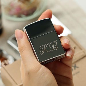 K&ouml;nnte beinhalten: Ein schwarzes und silbernes Feuerzeug mit den Initialen "K.B." in kursiver Schrift graviert. Das Feuerzeug wird in einer Hand gehalten und zeigt sein schlankes Design und die metallischen Akzente. Es ist ein klassischer Stil.