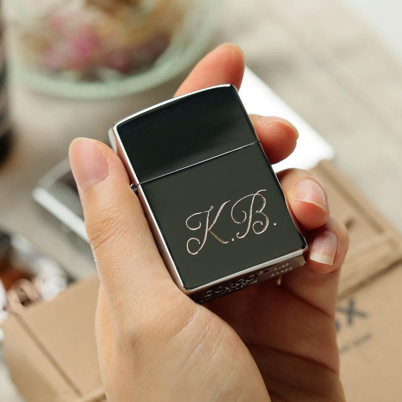 K&ouml;nnte beinhalten: Ein schwarzes und silbernes Feuerzeug mit den Initialen "K.B." in kursiver Schrift graviert. Das Feuerzeug wird in einer Hand gehalten und zeigt sein schlankes Design und die metallischen Akzente. Es ist ein klassischer Stil.