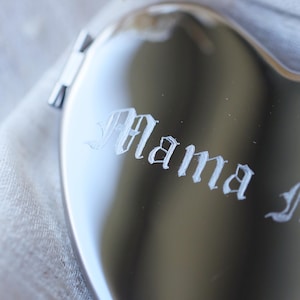 May include: A heart-shaped, silver-colored compact mirror with the word "Mama" etched in a decorative font. The mirror is partially open, revealing a glimpse of the interior. The background is a soft, neutral fabric.