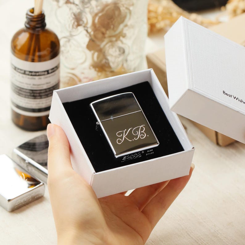 K&ouml;nnte beinhalten: Ein silbernes Zippo-Feuerzeug mit den Initialen "K.B." graviert, pr&auml;sentiert in einer wei&szlig;en Geschenkbox mit schwarzem Innenraum. Die Box wird in einer Hand gehalten.