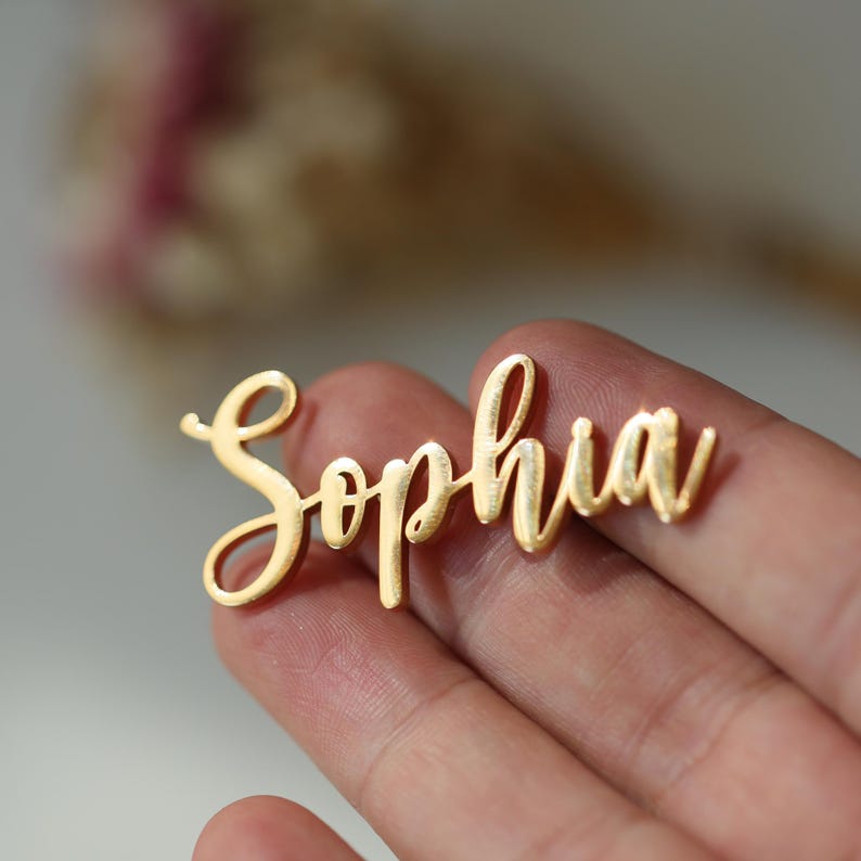 May include: A gold-colored metal nameplate in a cursive font that spells "Sophia". The nameplate is held in a hand, with a blurred background. The nameplate is likely used for jewelry or crafts.