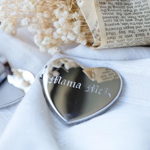 May include: A heart-shaped, silver-colored locket with the engraved text "Mama McR" in an elegant font. The locket is displayed on a white surface, with dried flowers and a piece of paper in the background.