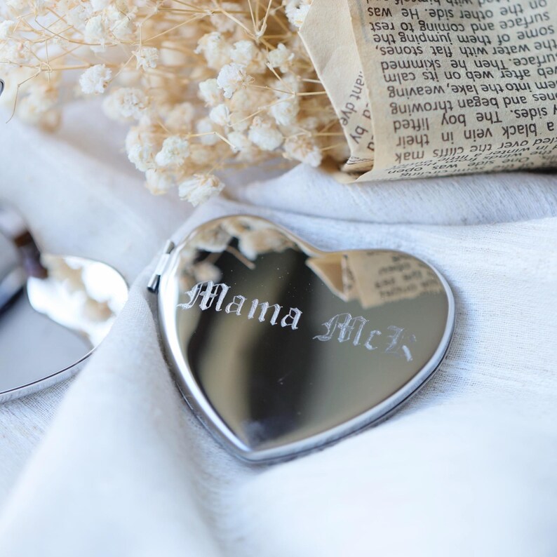 May include: A heart-shaped, silver-colored locket with the engraved text "Mama McR" in an elegant font. The locket is displayed on a white surface, with dried flowers and a piece of paper in the background.
