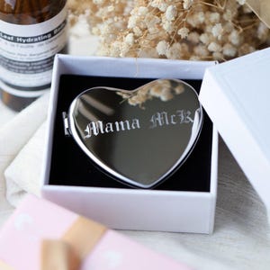 May include: A heart-shaped, silver compact mirror engraved with "Mama McK" in an elegant font, presented in a white gift box with a black interior. The mirror is surrounded by dried flowers, suggesting a gift or keepsake.