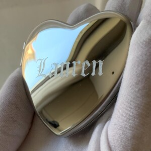 May include: A heart-shaped, silver-colored locket with the name "Lauren" engraved on the front. The locket is highly reflective, with a polished surface. The image shows the locket held in a gloved hand.