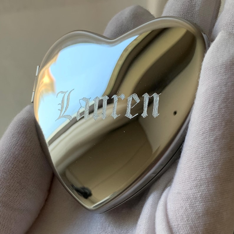 May include: A heart-shaped, silver-colored locket with the name "Lauren" engraved on the front. The locket is highly reflective, with a polished surface. The image shows the locket held in a gloved hand.