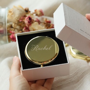 Puede incluir: Un espejo compacto redondo de color dorado con el nombre "Rachel" grabado. El espejo se presenta en una caja de regalo blanca con interior negro. La caja está parcialmente abierta, revelando el espejo.