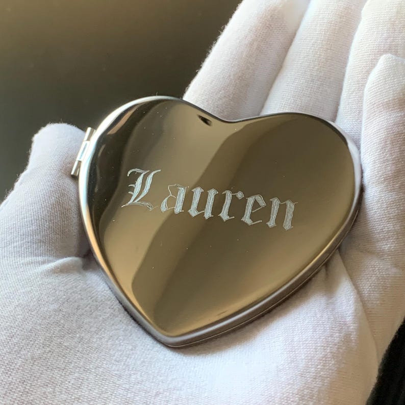 May include: A heart-shaped, silver compact mirror with the name "Lauren" engraved in an elegant font. The compact is held against a white background, showcasing its polished surface and intricate detailing. A personalized gift.