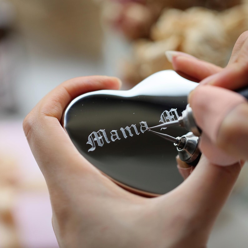 May include: A heart-shaped, silver-colored keepsake is being engraved with the word "Mama" in an elegant script. The engraving tool is held by a person, adding a personalized touch to the item. The background is blurred, focusing attention on the heart.