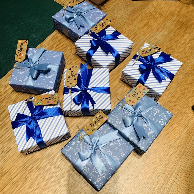May include: An assortment of wrapped gifts with blue ribbons and name tags. The gifts are wrapped in various patterned papers, including stripes and whale designs. Each gift is tied with a vibrant blue ribbon and a personalized tag.