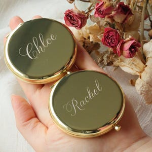 May include: Two gold-colored compact mirrors, one inscribed with "Chloe" and the other with "Rachel" in elegant script. The mirrors are round with a shiny finish. The background includes dried roses.