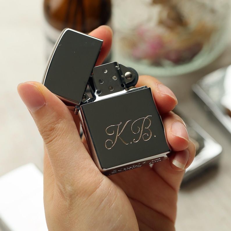 K&ouml;nnte beinhalten: Ein silberfarbenes Feuerzeug mit ge&ouml;ffnetem Deckel, gehalten in einer Hand. Das Feuerzeug hat die Initialen "K.B." auf der Vorderseite eingraviert. Der Hintergrund ist unscharf und zeigt andere Objekte.