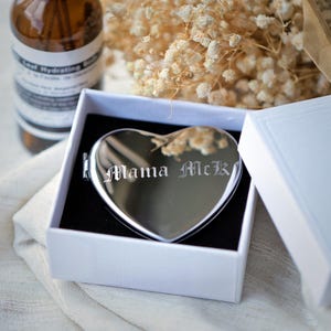May include: A heart-shaped, silver compact mirror with the engraved text "Mama McK" inside a white gift box with a black interior. The mirror is reflective, and the box is open, revealing the mirror. A brown bottle and dried flowers are in the background.